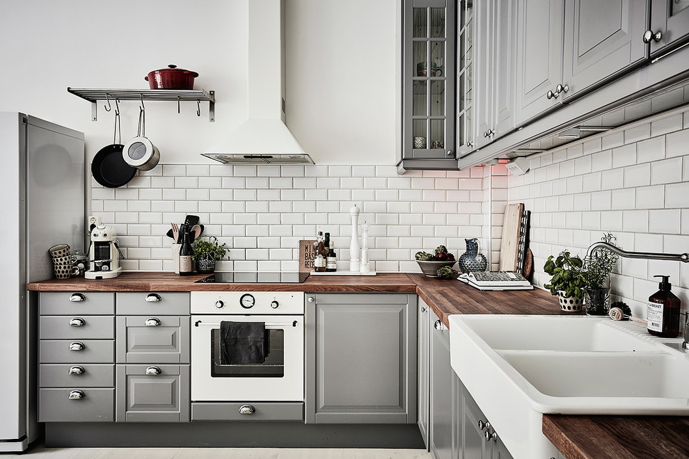 Idéer för mellanstora minimalistiska l-kök, med en rustik diskho, luckor med upphöjd panel, grå skåp, träbänkskiva, vitt stänkskydd, stänkskydd i tunnelbanekakel, vita vitvaror och ljust trägolv