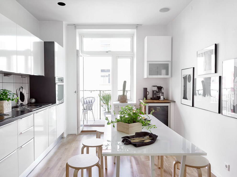 Medium sized scandi single-wall kitchen/diner in Gothenburg with a built-in sink, flat-panel cabinets, white cabinets, white splashback, stainless steel appliances, light hardwood flooring, granite worktops and no island.