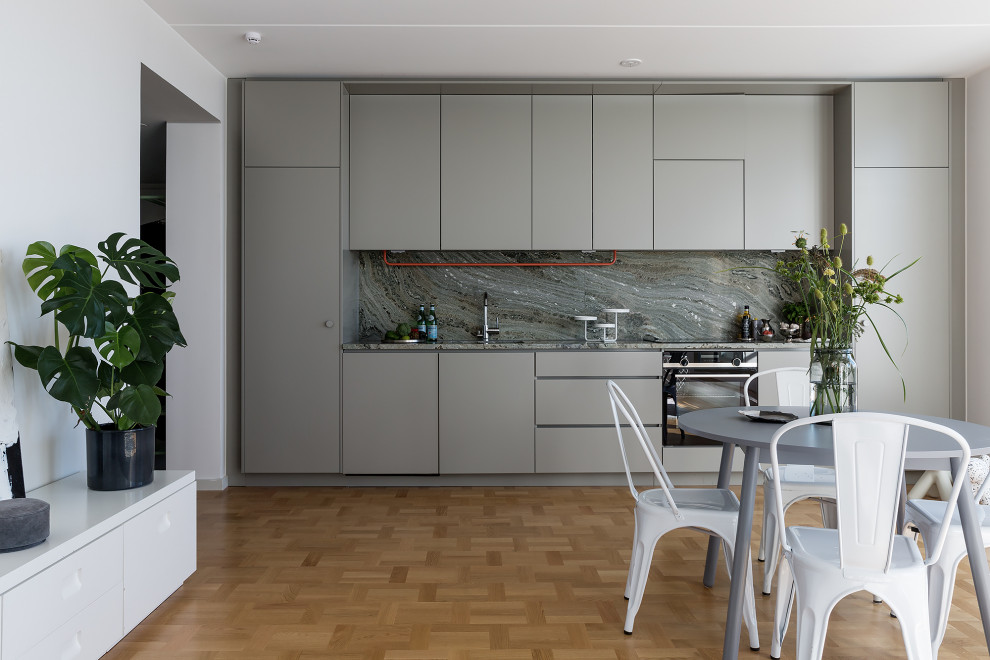 Minimalistisk inredning av ett grå linjärt grått kök med öppen planlösning, med släta luckor, grå skåp, grått stänkskydd, stänkskydd i sten, rostfria vitvaror, mellanmörkt trägolv och brunt golv