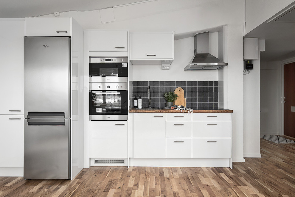 Idéer för att renovera ett mellanstort skandinaviskt linjärt kök med öppen planlösning, med en enkel diskho, släta luckor, vita skåp, träbänkskiva, svart stänkskydd, stänkskydd i keramik, rostfria vitvaror och ljust trägolv