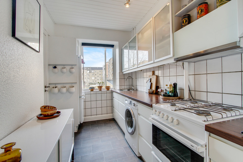 Foto på ett avskilt, litet funkis linjärt kök, med en nedsänkt diskho, släta luckor, vita skåp, vitt stänkskydd, vita vitvaror, träbänkskiva, stänkskydd i porslinskakel och klinkergolv i keramik