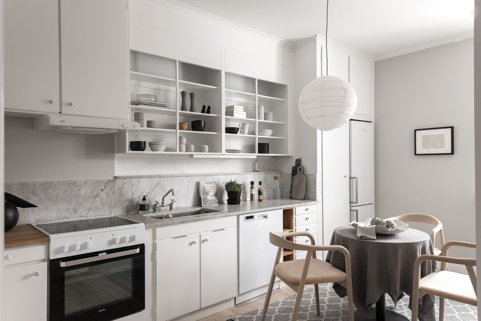 Skandinavisk inredning av ett grå linjärt grått kök och matrum, med en undermonterad diskho, släta luckor, vita skåp, grått stänkskydd, stänkskydd i sten, vita vitvaror, ljust trägolv och beiget golv