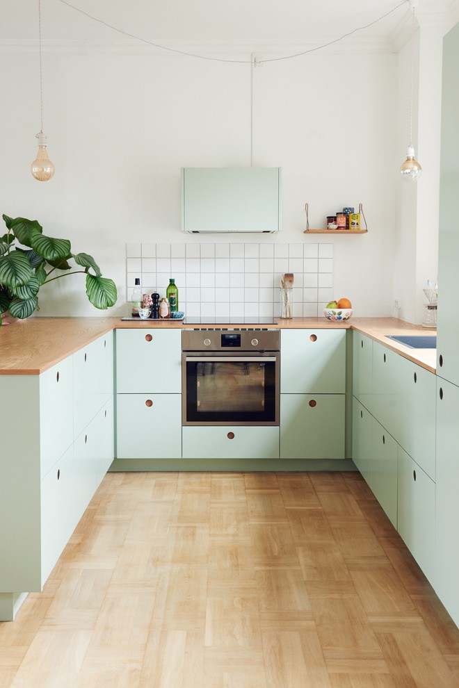 コペンハーゲンにある北欧スタイルのおしゃれなペニンシュラキッチン (ドロップインシンク、フラットパネル扉のキャビネット、緑のキャビネット、木材カウンター、白いキッチンパネル、シルバーの調理設備、無垢フローリング) の写真