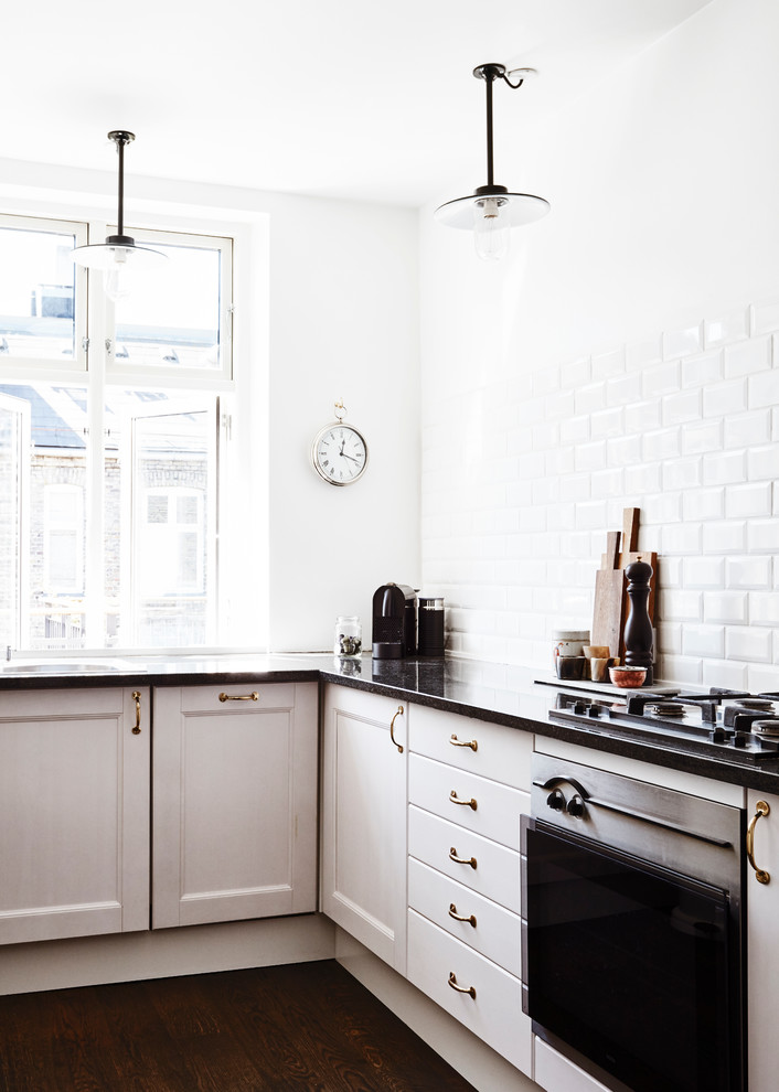 Inredning av ett minimalistiskt l-kök, med luckor med infälld panel, vita skåp, vitt stänkskydd, rostfria vitvaror, mörkt trägolv och stänkskydd i keramik