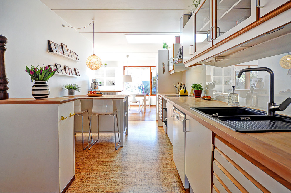 Exempel på ett mellanstort modernt linjärt kök med öppen planlösning, med en nedsänkt diskho, släta luckor, vita skåp, träbänkskiva, rostfria vitvaror, en halv köksö, grått stänkskydd, glaspanel som stänkskydd och korkgolv