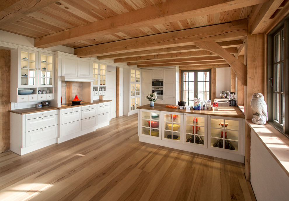 Bild på ett stort lantligt kök, med luckor med glaspanel, vita skåp, ljust trägolv, en halv köksö, en nedsänkt diskho, träbänkskiva, grått stänkskydd, stänkskydd i cementkakel och rostfria vitvaror