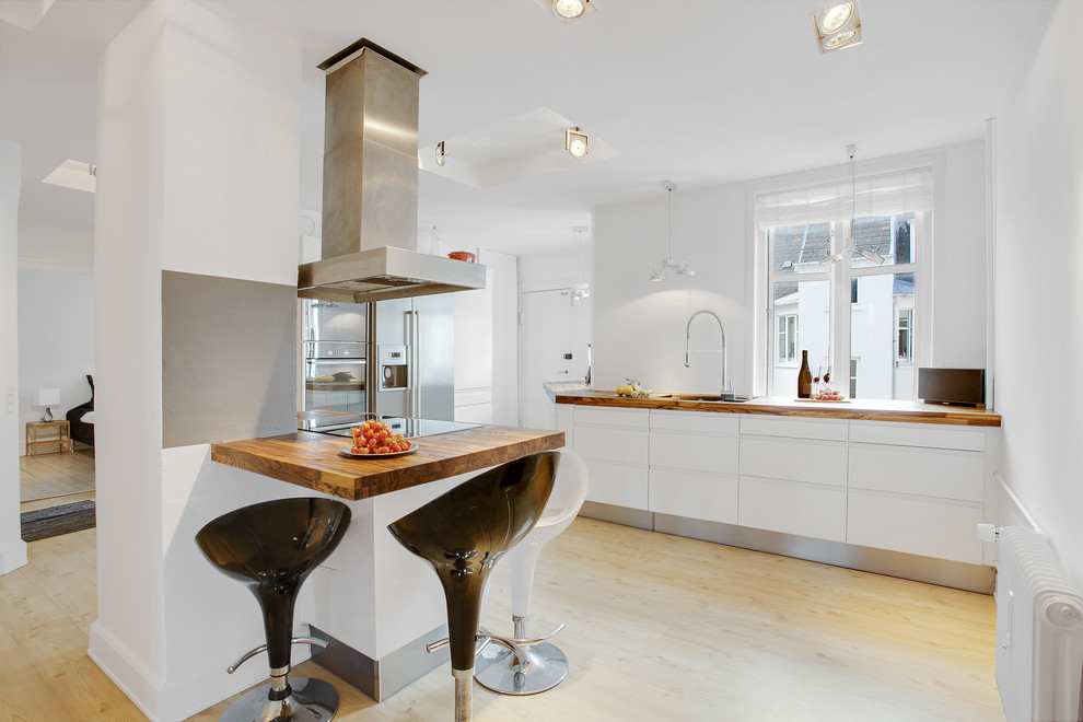 Medium sized scandi l-shaped enclosed kitchen in Miami with a submerged sink, flat-panel cabinets, white cabinets, wood worktops, grey splashback, stainless steel appliances, light hardwood flooring, an island and glass sheet splashback.