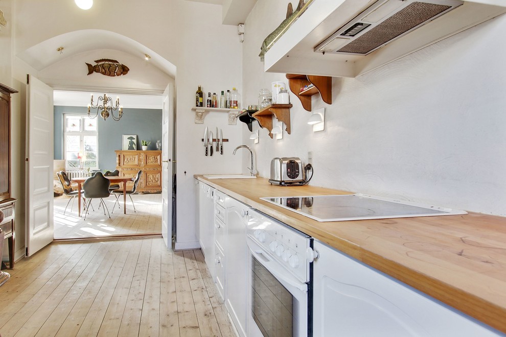 Nordisk inredning av ett avskilt, mellanstort linjärt kök, med en nedsänkt diskho, luckor med upphöjd panel, vita skåp, träbänkskiva, vita vitvaror och ljust trägolv