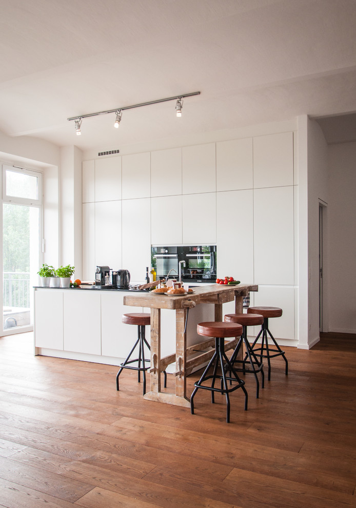 Small contemporary galley open plan kitchen in Berlin with flat-panel cabinets, white cabinets, an island, medium hardwood flooring, white splashback and black appliances.