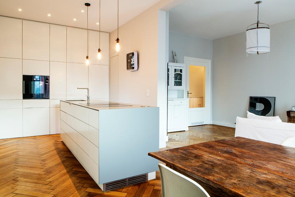 Example of a mid-sized trendy single-wall medium tone wood floor and brown floor open concept kitchen design in Munich with flat-panel cabinets, white cabinets, an island, an integrated sink, solid surface countertops, white backsplash, limestone backsplash and stainless steel appliances