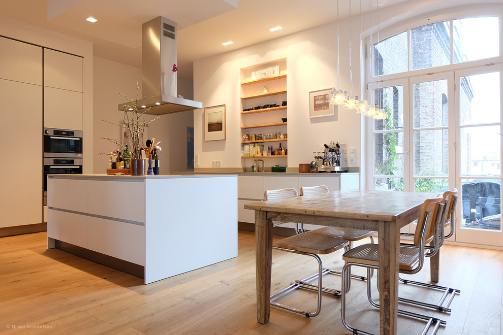 Offene, Große Moderne Küche mit flächenbündigen Schrankfronten, weißen Schränken, Kücheninsel, Küchengeräten aus Edelstahl, Küchenrückwand in Weiß und braunem Holzboden in Köln