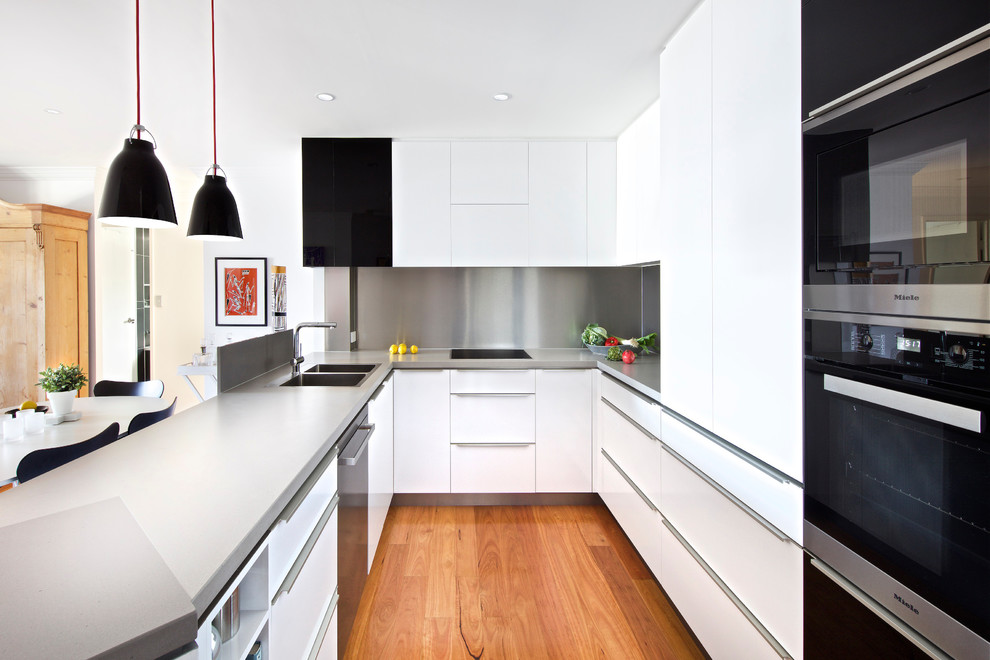 На фото: п-образная кухня в современном стиле с двойной мойкой, плоскими фасадами, белыми фасадами, фартуком цвета металлик, черной техникой, паркетным полом среднего тона, полуостровом, коричневым полом и серой столешницей