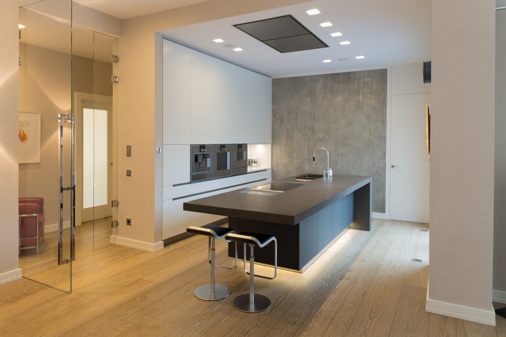This is an example of a large modern galley open plan kitchen in Munich with an integrated sink, flat-panel cabinets, white cabinets, medium hardwood flooring, an island, brown floors, black worktops, soapstone worktops, white splashback, glass sheet splashback and stainless steel appliances.