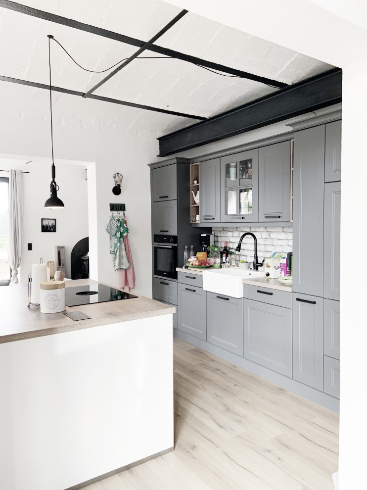 Inspiration for a large contemporary single-wall open plan kitchen in Berlin with a belfast sink, raised-panel cabinets, grey cabinets, soapstone worktops, white splashback, brick splashback, light hardwood flooring, multiple islands, beige worktops and a vaulted ceiling.