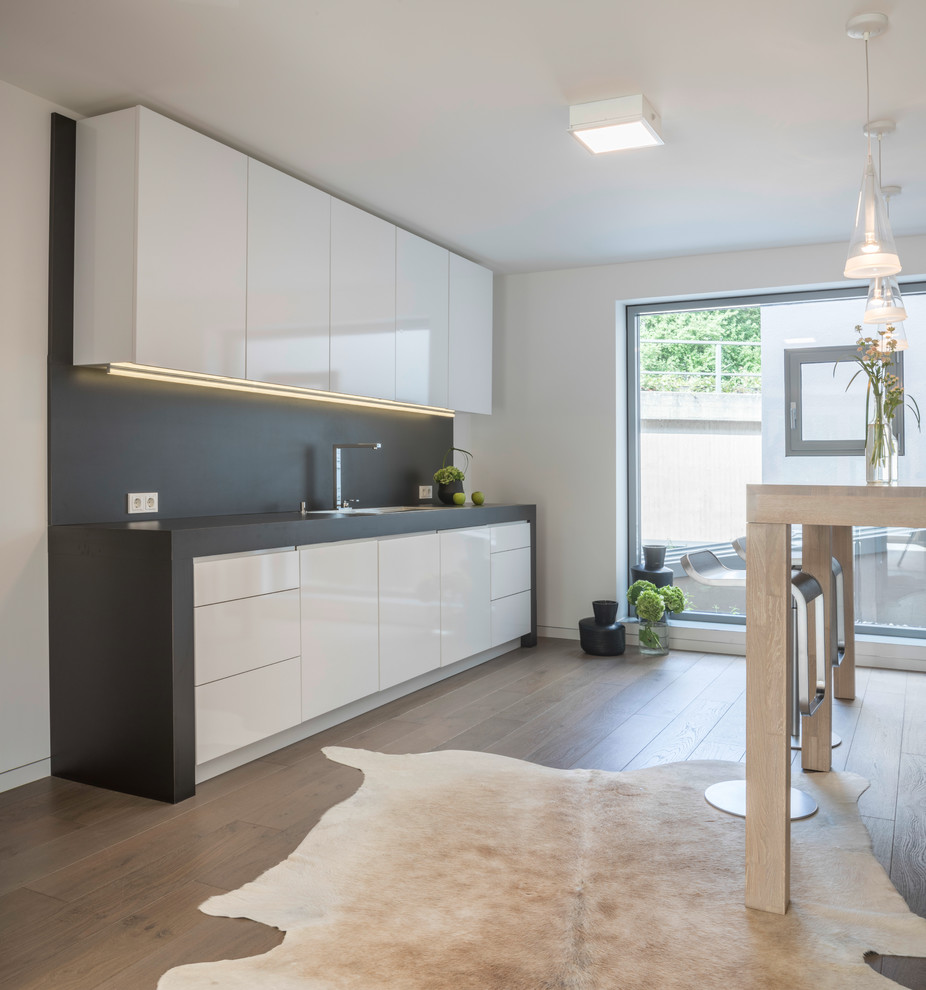 Cette image montre une petite cuisine ouverte linéaire minimaliste avec un évier posé, un placard à porte plane, des portes de placard blanches, une crédence grise, un sol en bois brun, aucun îlot et un sol marron.