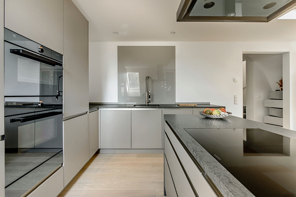 Design ideas for a contemporary l-shaped open plan kitchen in Munich with a built-in sink, flat-panel cabinets, grey cabinets, soapstone worktops, glass sheet splashback, black appliances, light hardwood flooring and an island.