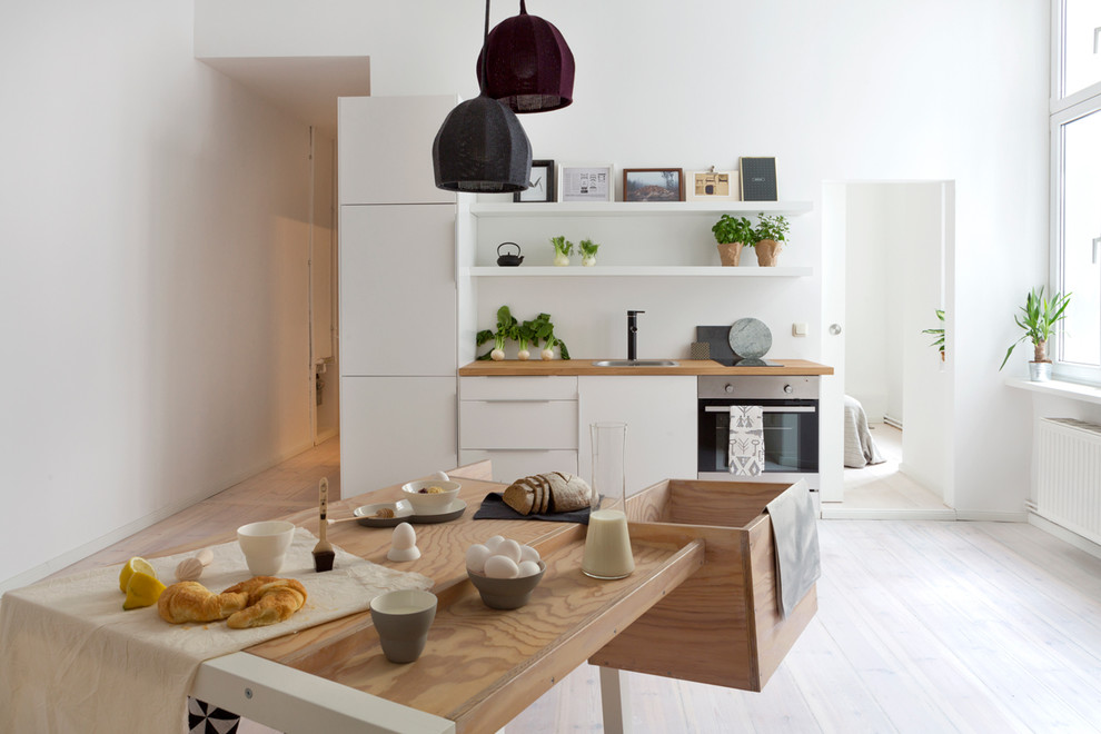 ベルリンにある小さな北欧スタイルのおしゃれなキッチン (ドロップインシンク、フラットパネル扉のキャビネット、白いキャビネット、木材カウンター、白いキッチンパネル、パネルと同色の調理設備、淡色無垢フローリング) の写真