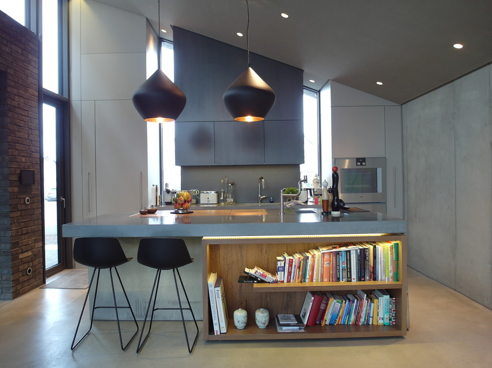 Cette image montre une grande cuisine linéaire minimaliste avec un placard à porte plane, des portes de placard grises, un plan de travail en béton, une crédence grise, un électroménager en acier inoxydable, sol en béton ciré et îlot.