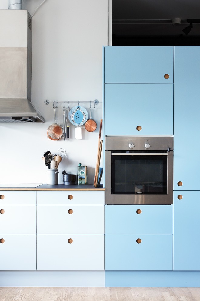 Diseño de cocinas en L escandinava de tamaño medio cerrada sin isla con puertas de armario azules, suelo de madera clara, electrodomésticos de acero inoxidable, armarios con paneles lisos y encimera de madera