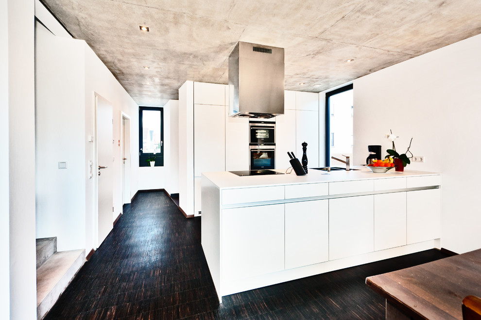 На фото: кухня-гостиная среднего размера в современном стиле с монолитной мойкой, плоскими фасадами, белыми фасадами, темным паркетным полом, черной техникой и полуостровом