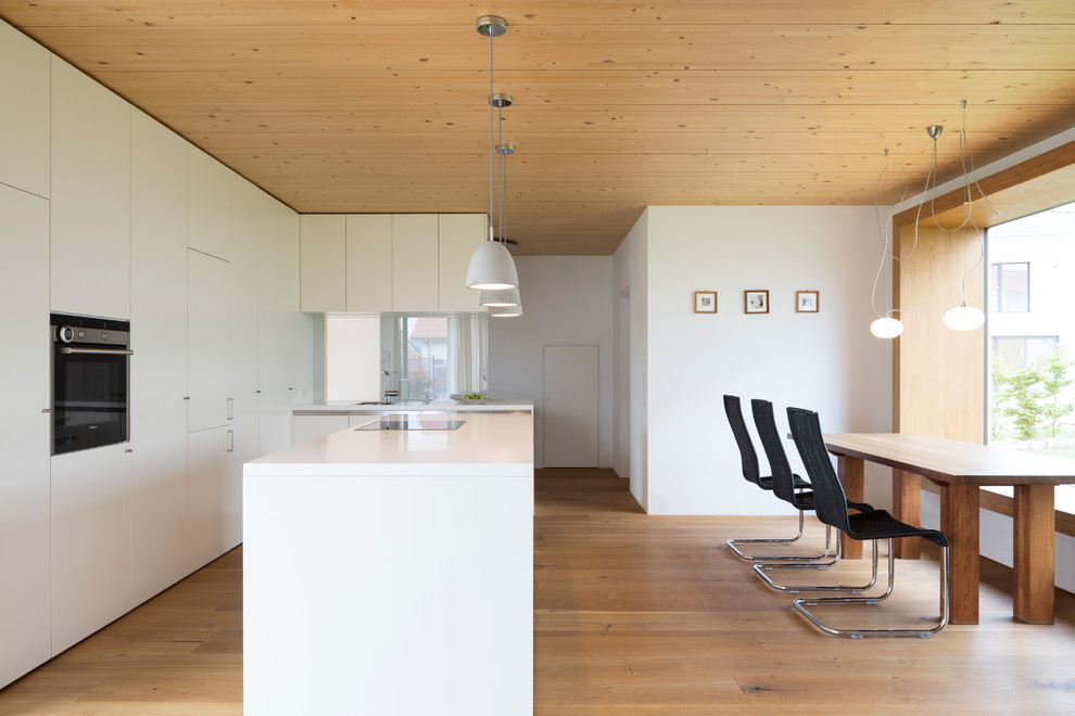 Große Moderne Wohnküche in L-Form mit flächenbündigen Schrankfronten, weißen Schränken, Elektrogeräten mit Frontblende, hellem Holzboden, Kücheninsel und Waschbecken in Sonstige