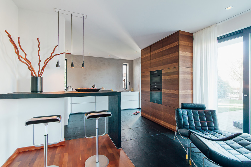 Inspiration for a medium sized contemporary l-shaped open plan kitchen in Frankfurt with a submerged sink, flat-panel cabinets, composite countertops, grey splashback, black appliances, slate flooring, black floors, white worktops, medium wood cabinets and a breakfast bar.