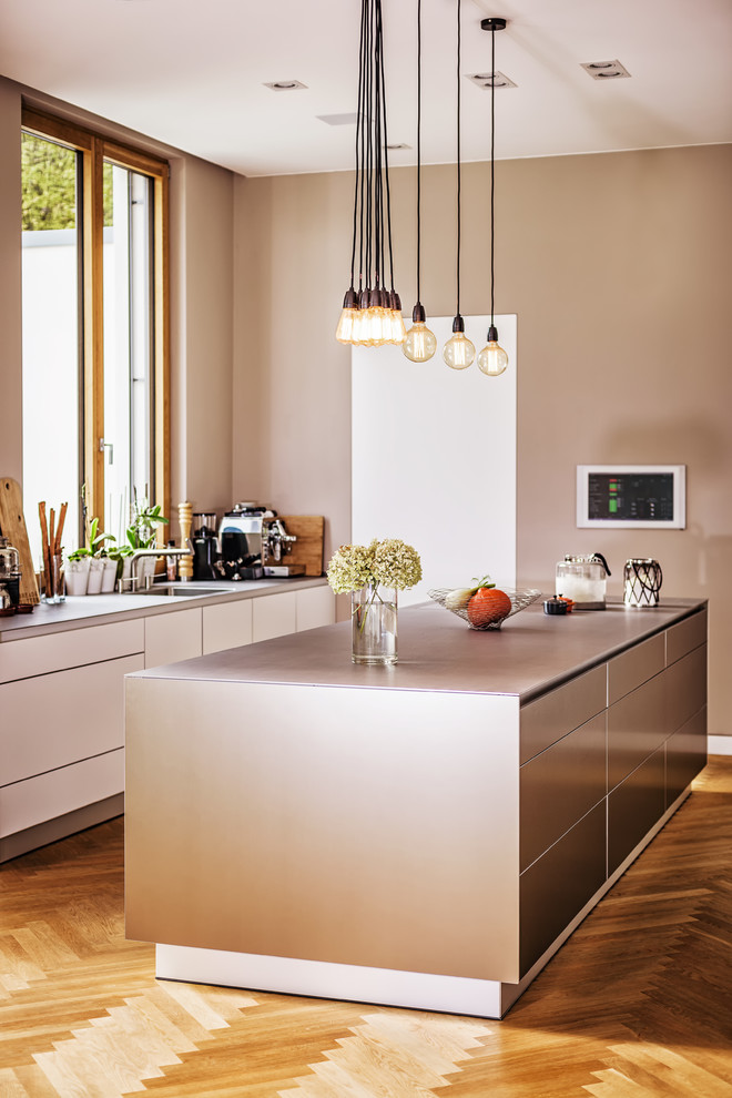 Inredning av ett modernt stort grå grått kök, med en integrerad diskho, släta luckor, beige skåp, bänkskiva i rostfritt stål, rostfria vitvaror, mellanmörkt trägolv, en köksö och brunt golv