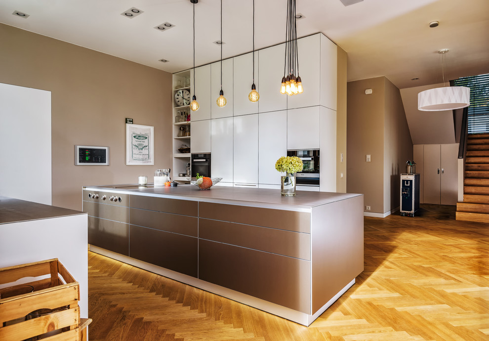 This is an example of a large contemporary galley open plan kitchen in Berlin with an integrated sink, flat-panel cabinets, beige cabinets, stainless steel worktops, stainless steel appliances, medium hardwood flooring, an island, brown floors and grey worktops.