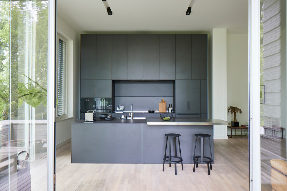 Modern inredning av ett stort svart linjärt svart kök och matrum, med släta luckor, svarta skåp, svart stänkskydd, svarta vitvaror, ljust trägolv, en köksö, beiget golv, en enkel diskho och bänkskiva i betong
