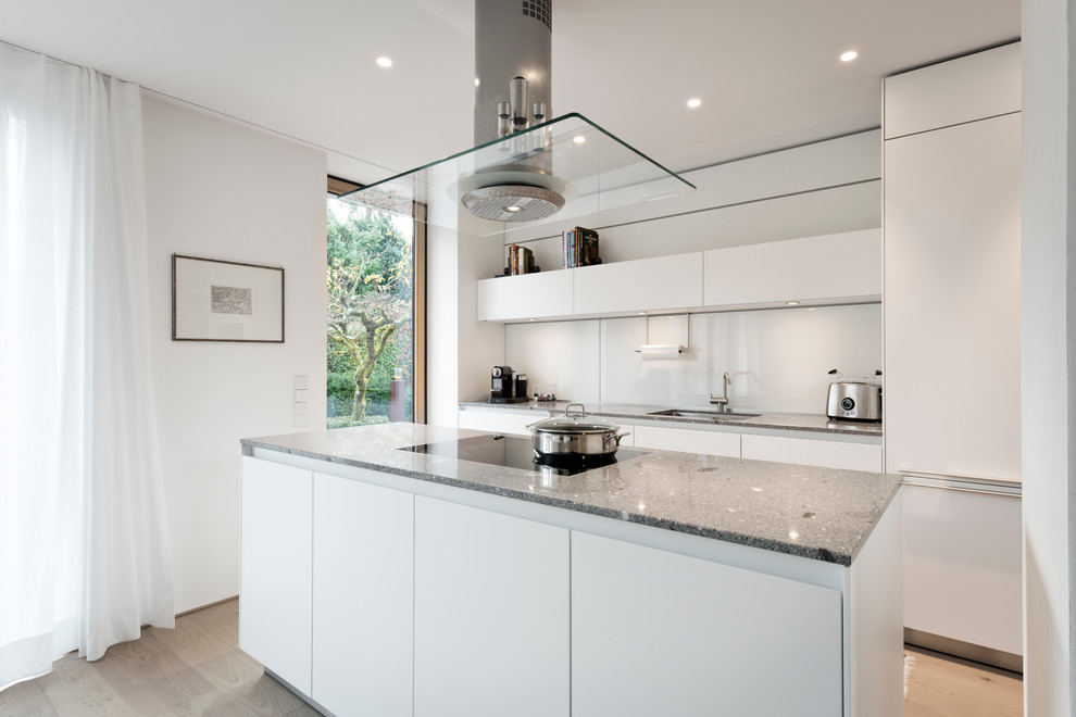 Modern inredning av ett avskilt, mellanstort kök, med en undermonterad diskho, släta luckor, vita skåp, vitt stänkskydd, glaspanel som stänkskydd, mellanmörkt trägolv och en köksö