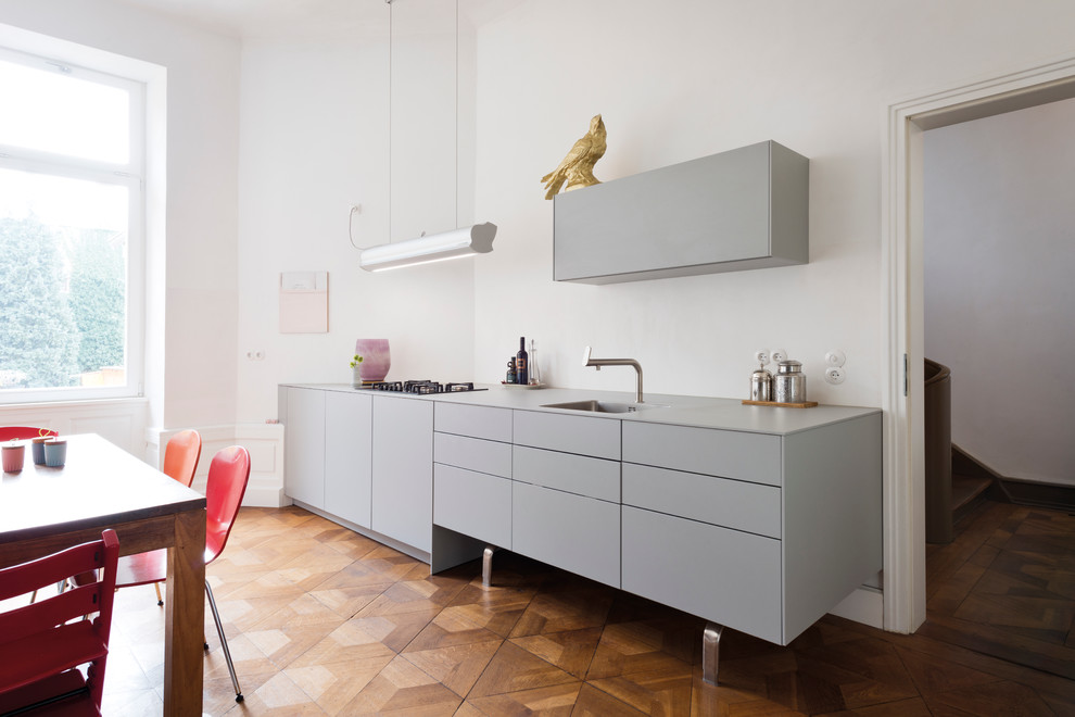 Einzeilige, Mittelgroße Moderne Wohnküche ohne Insel mit Unterbauwaschbecken, flächenbündigen Schrankfronten, grauen Schränken, Küchenrückwand in Weiß und braunem Holzboden in Berlin
