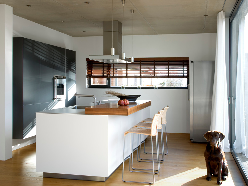 Exempel på ett mellanstort modernt kök med öppen planlösning, med en integrerad diskho, bänkskiva i rostfritt stål, rostfria vitvaror, mellanmörkt trägolv, en köksö, släta luckor och grå skåp