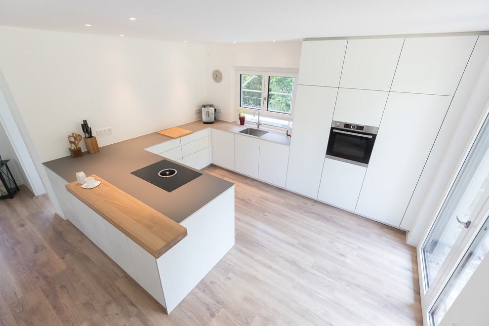 Design ideas for a large modern l-shaped open plan kitchen in Stuttgart with a built-in sink, flat-panel cabinets, white cabinets, laminate countertops, white splashback, wood splashback, stainless steel appliances, laminate floors, a breakfast bar, brown floors and grey worktops.