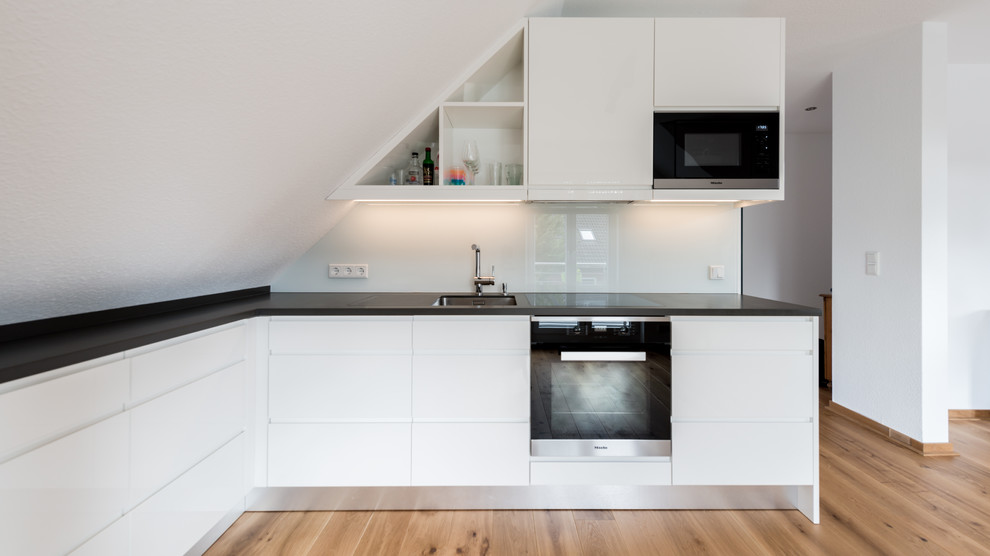 Bild på ett mellanstort funkis kök, med en nedsänkt diskho, släta luckor, vita skåp, vitt stänkskydd, glaspanel som stänkskydd, svarta vitvaror, mellanmörkt trägolv, brunt golv och bänkskiva i koppar