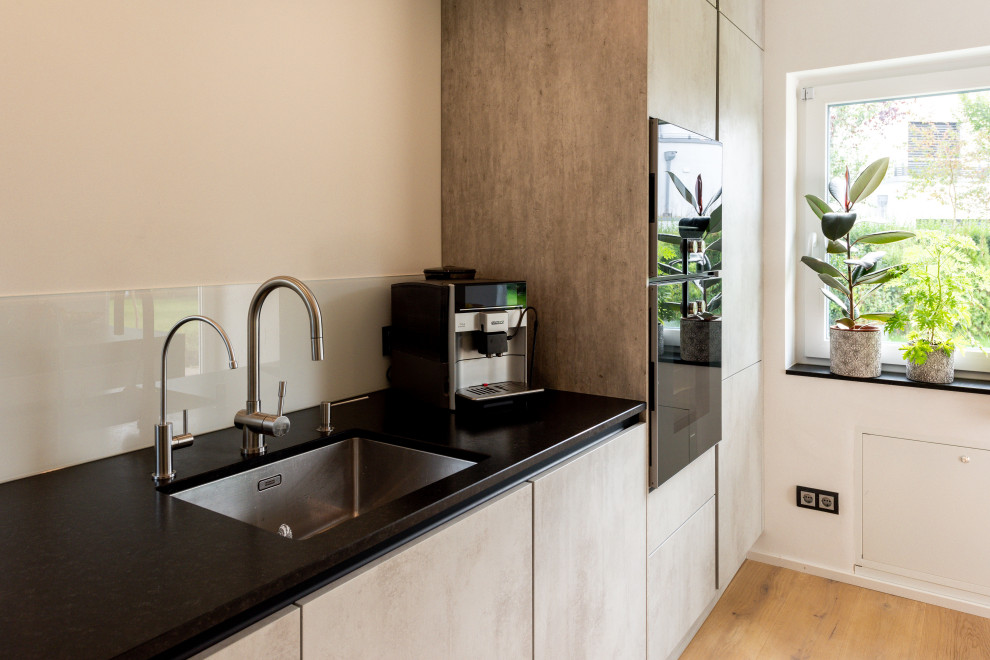 Zweizeilige Moderne Küche mit flächenbündigen Schrankfronten, grauen Schränken, Granit-Arbeitsplatte, schwarzen Elektrogeräten, braunem Holzboden, Kücheninsel und schwarzer Arbeitsplatte in München