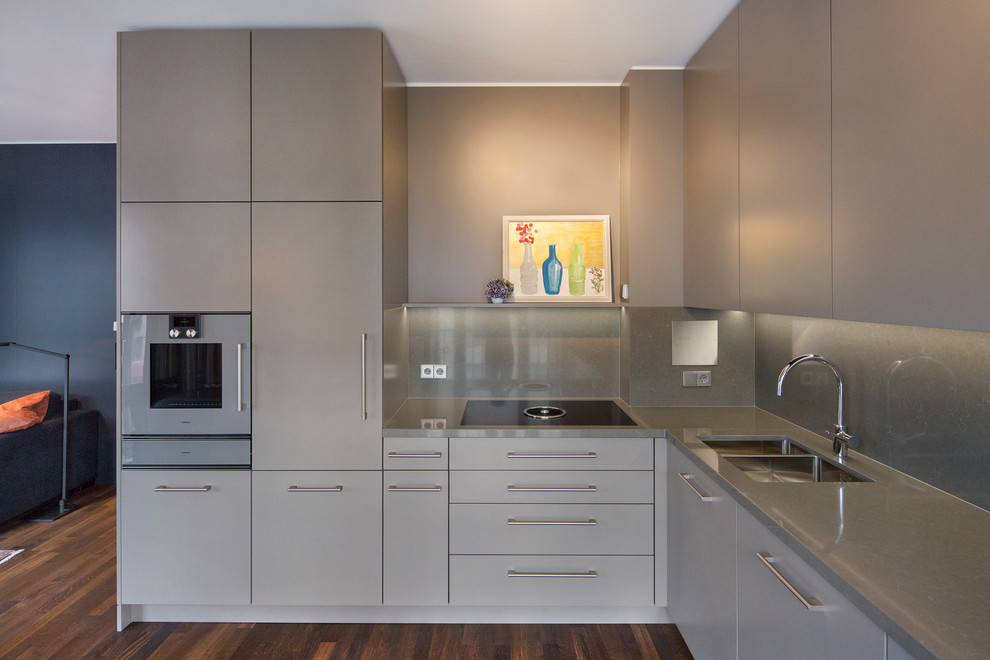 На фото: угловая кухня-гостиная среднего размера в современном стиле с накладной мойкой, серыми фасадами, паркетным полом среднего тона и гранитной столешницей с