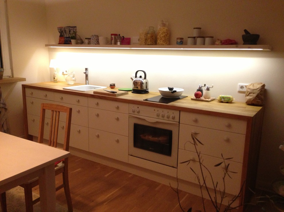 Bild på ett mellanstort skandinaviskt linjärt kök och matrum, med en nedsänkt diskho, släta luckor, vita skåp, träbänkskiva, beige stänkskydd, vita vitvaror, ljust trägolv och brunt golv
