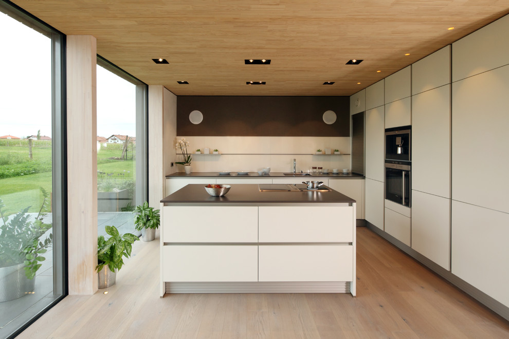 Large contemporary l-shaped open plan kitchen in Munich with flat-panel cabinets, white cabinets, stainless steel appliances, light hardwood flooring, an island, an integrated sink, white splashback, brown floors and black worktops.