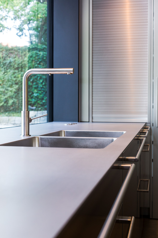 Inredning av ett modernt stort grå linjärt grått kök med öppen planlösning, med en integrerad diskho, släta luckor, skåp i rostfritt stål, bänkskiva i rostfritt stål, stänkskydd med metallisk yta, rostfria vitvaror och en köksö
