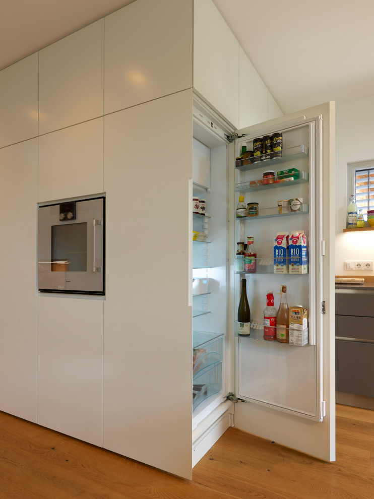 This is an example of a large contemporary galley open plan kitchen in Munich with an integrated sink, flat-panel cabinets, white cabinets, concrete worktops, white splashback, wood splashback, stainless steel appliances, medium hardwood flooring, an island, brown floors and grey worktops.