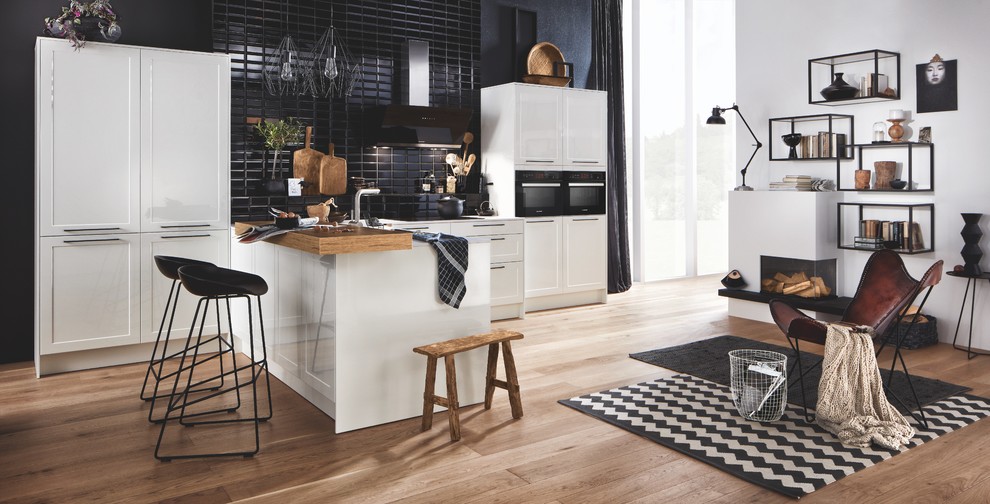 Bild på ett litet funkis vit linjärt vitt kök och matrum, med skåp i shakerstil, vita skåp, svart stänkskydd, stänkskydd i tunnelbanekakel, svarta vitvaror, mellanmörkt trägolv, en halv köksö, brunt golv och bänkskiva i glas