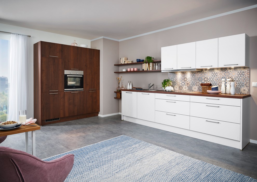 Modern inredning av ett mellanstort brun brunt kök, med en nedsänkt diskho, släta luckor, träbänkskiva, flerfärgad stänkskydd, svarta vitvaror, grått golv, vita skåp och glaspanel som stänkskydd