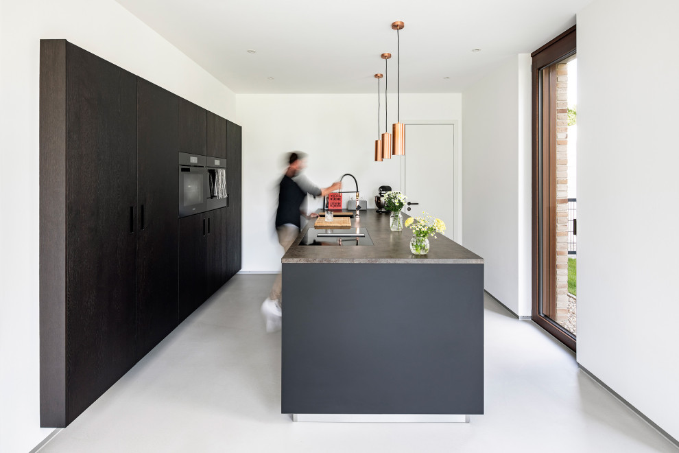 Aménagement d'une cuisine parallèle moderne avec un placard à porte plane, des portes de placard noires, un électroménager noir, sol en béton ciré, îlot, un sol gris et un plan de travail gris.