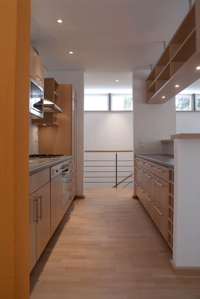 На фото: параллельная кухня-гостиная среднего размера в современном стиле с плоскими фасадами, фасадами цвета дерева среднего тона, белым фартуком и техникой из нержавеющей стали