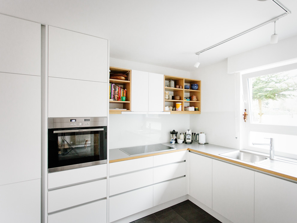 Foto på ett litet skandinaviskt l-kök, med en enkel diskho, släta luckor, vita skåp, vitt stänkskydd, glaspanel som stänkskydd, rostfria vitvaror och svart golv