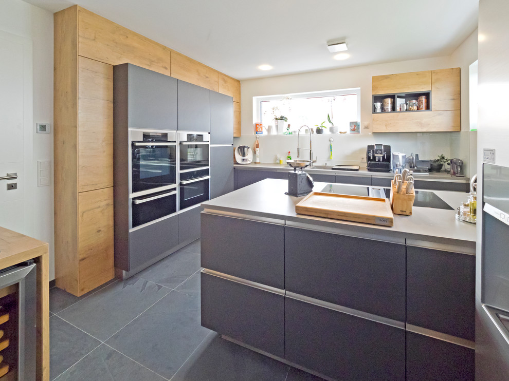 Mittelgroße Moderne Küche in L-Form mit flächenbündigen Schrankfronten, grauen Schränken, Glasrückwand, Küchengeräten aus Edelstahl, Kücheninsel und grauem Boden in Nürnberg