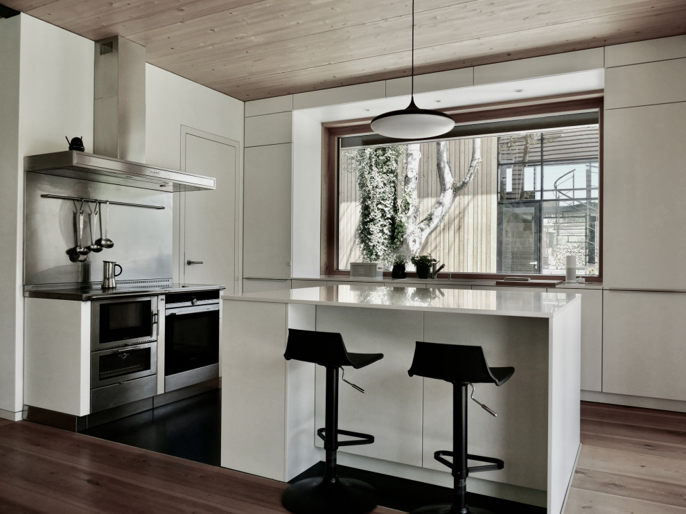 Modern inredning av ett vit vitt l-kök, med släta luckor, vita skåp, rostfria vitvaror, mellanmörkt trägolv, en köksö och brunt golv