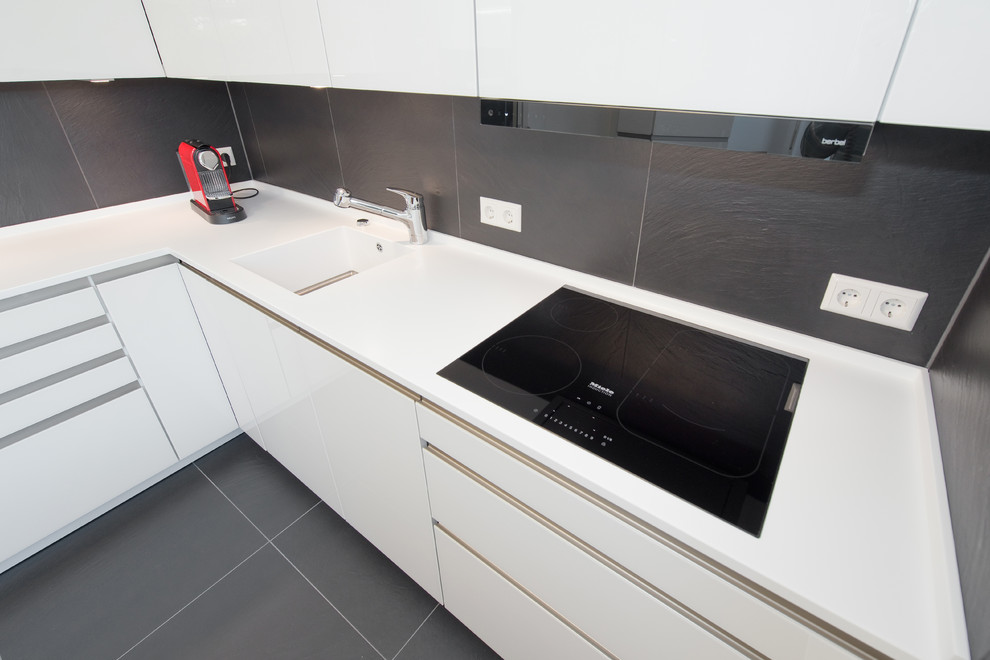 Kleine Moderne Wohnküche ohne Insel in L-Form mit integriertem Waschbecken, Glasfronten, weißen Schränken, Mineralwerkstoff-Arbeitsplatte, Küchenrückwand in Schwarz, schwarzen Elektrogeräten und Schieferboden in Stuttgart