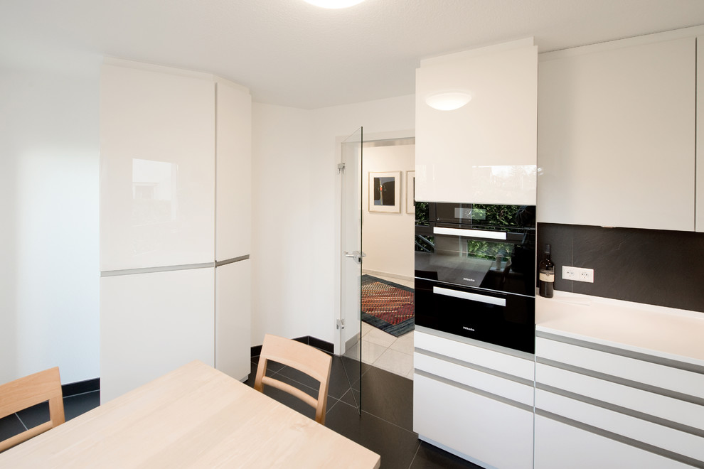 Small contemporary l-shaped kitchen/diner in Stuttgart with an integrated sink, glass-front cabinets, white cabinets, composite countertops, black splashback, black appliances, slate flooring and no island.