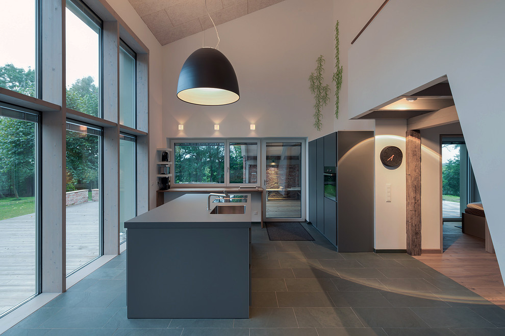 Cette photo montre une grande cuisine ouverte tendance avec un évier intégré, un placard à porte plane, des portes de placard grises, un électroménager noir, un sol en ardoise et îlot.